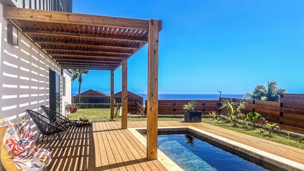 A wooden deck with a pool, outdoor seating, and a view of the ocean.