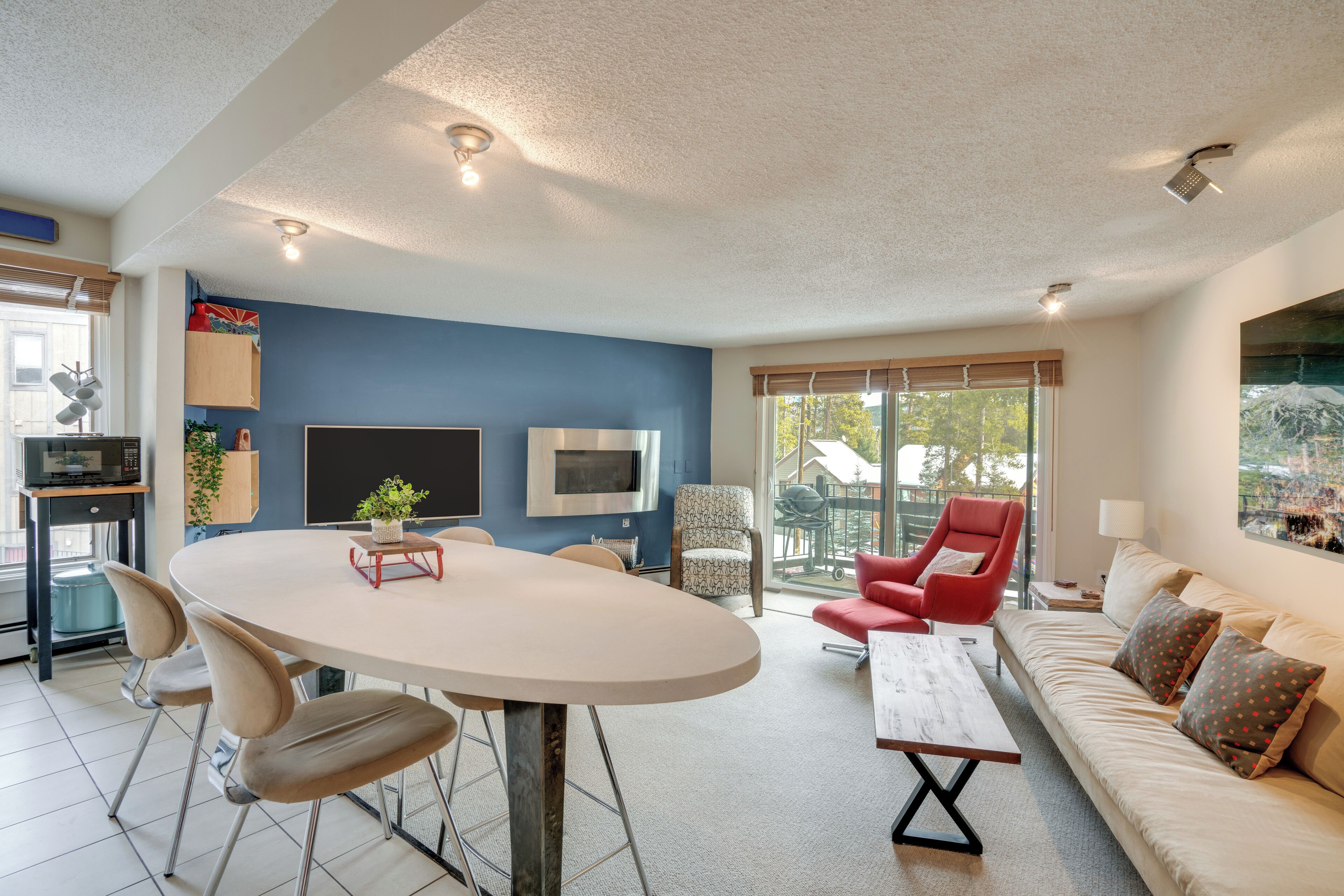 Een moderne woonkamer met een ronde tafel, twee stoelen, een bank en een rode fauteuil. Een televisie is aan de muur bevestigd, er is een raam met jaloezieën en een deur naar buiten.