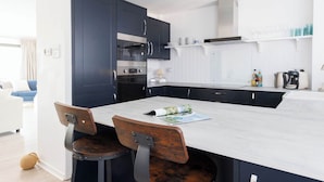 A modern kitchen with dark cabinets, a marble island, and a dining area with wooden chairs. - Beautifully Refurbished 4-Bed Beachside Retreat (Selsey)