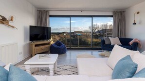 A modern living room with a large TV, a sofa, a coffee table, and a view through large windows. - Beautifully Refurbished 4-Bed Beachside Retreat (Selsey)