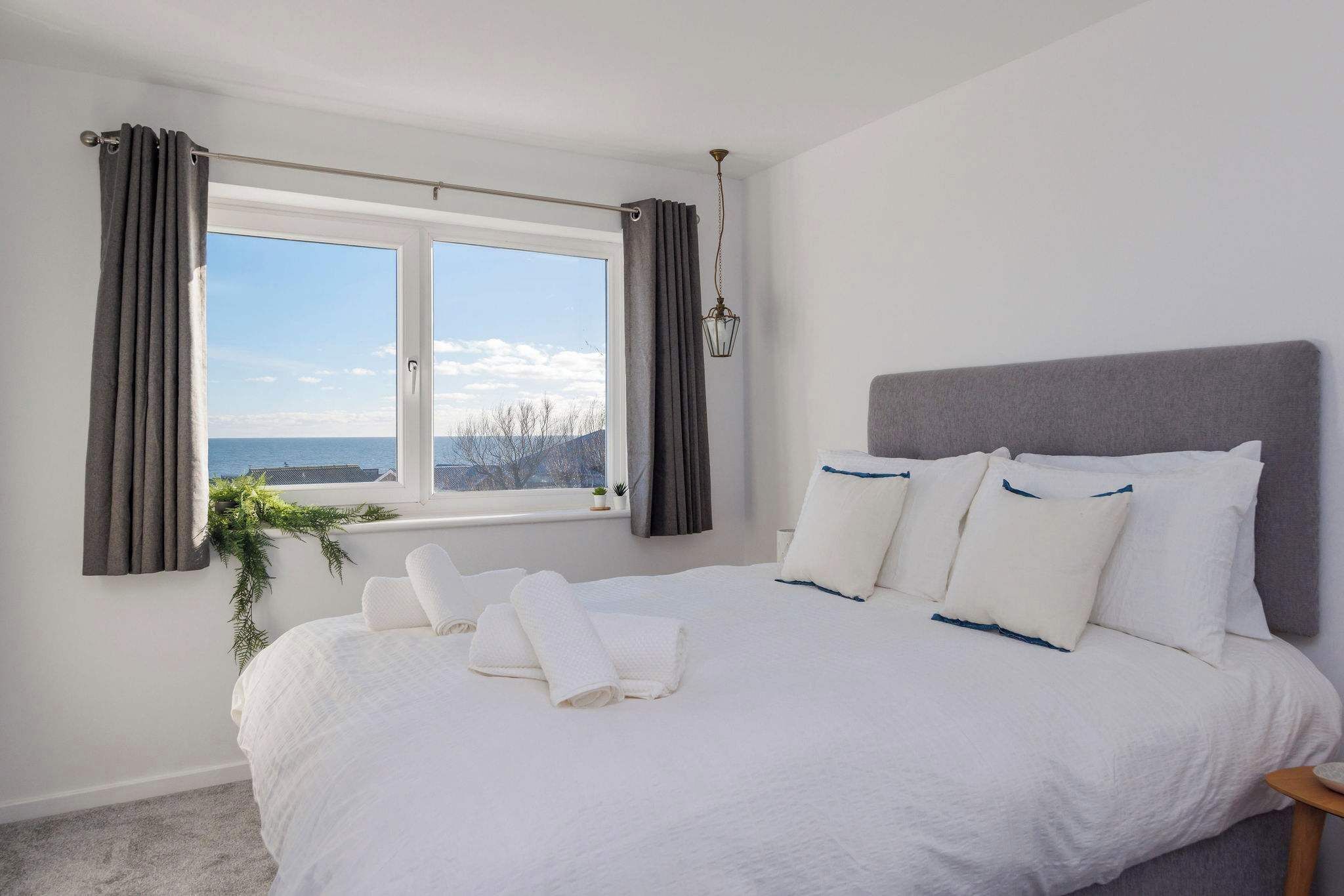 A neatly made bed with white linens and pillows, a window with curtains, and a view of the sea.
