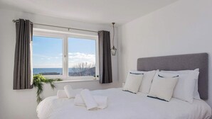 A neatly made bed with white linens and pillows, a window with curtains, and a view of the sea. - Beautifully Refurbished 4-Bed Beachside Retreat (Selsey)