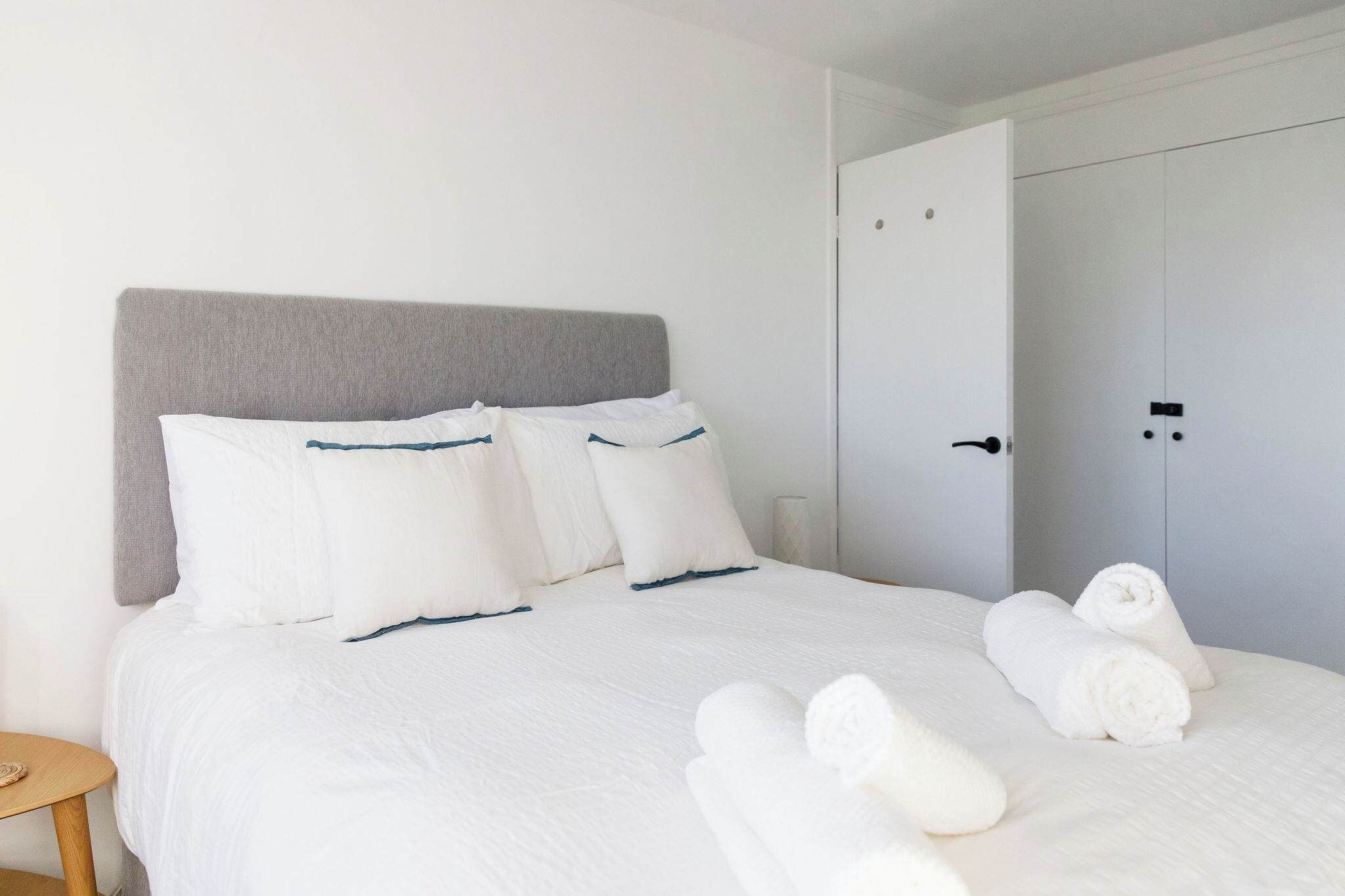 A neatly made bed with white linens and a grey headboard, a wooden nightstand, and a closed door in the background.
