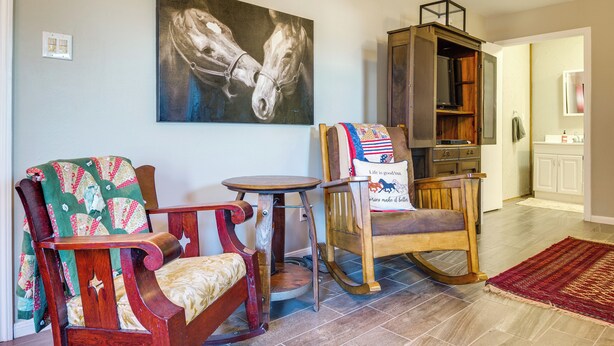 Een woonkamer met een schommelstoel, een houten tafel en een schilderij van paarden aan de muur.