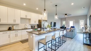 Een moderne keuken met witte kasten, een groot eiland met barkrukken, roestvrijstalen apparatuur, hanglampen en een zithoek met een bank en televisie.