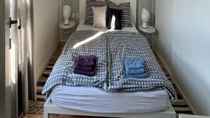 Una cama pequeña y bien hecha con una colcha estampada y almohadas, una mesita de noche con una lámpara, y una ventana con cortinas.