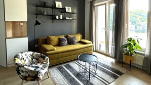 Una sala moderna con un sofá color mostaza, una mesa redonda negra y un sillón estampado con motivos florales. Hay estanterías con objetos decorativos y un cartel con la palabra 'Welcome' en la pared.