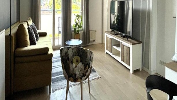 Una sala de estar con un sofá, un sillón floral, un televisor sobre un mueble de madera y un balcón con cortinas.