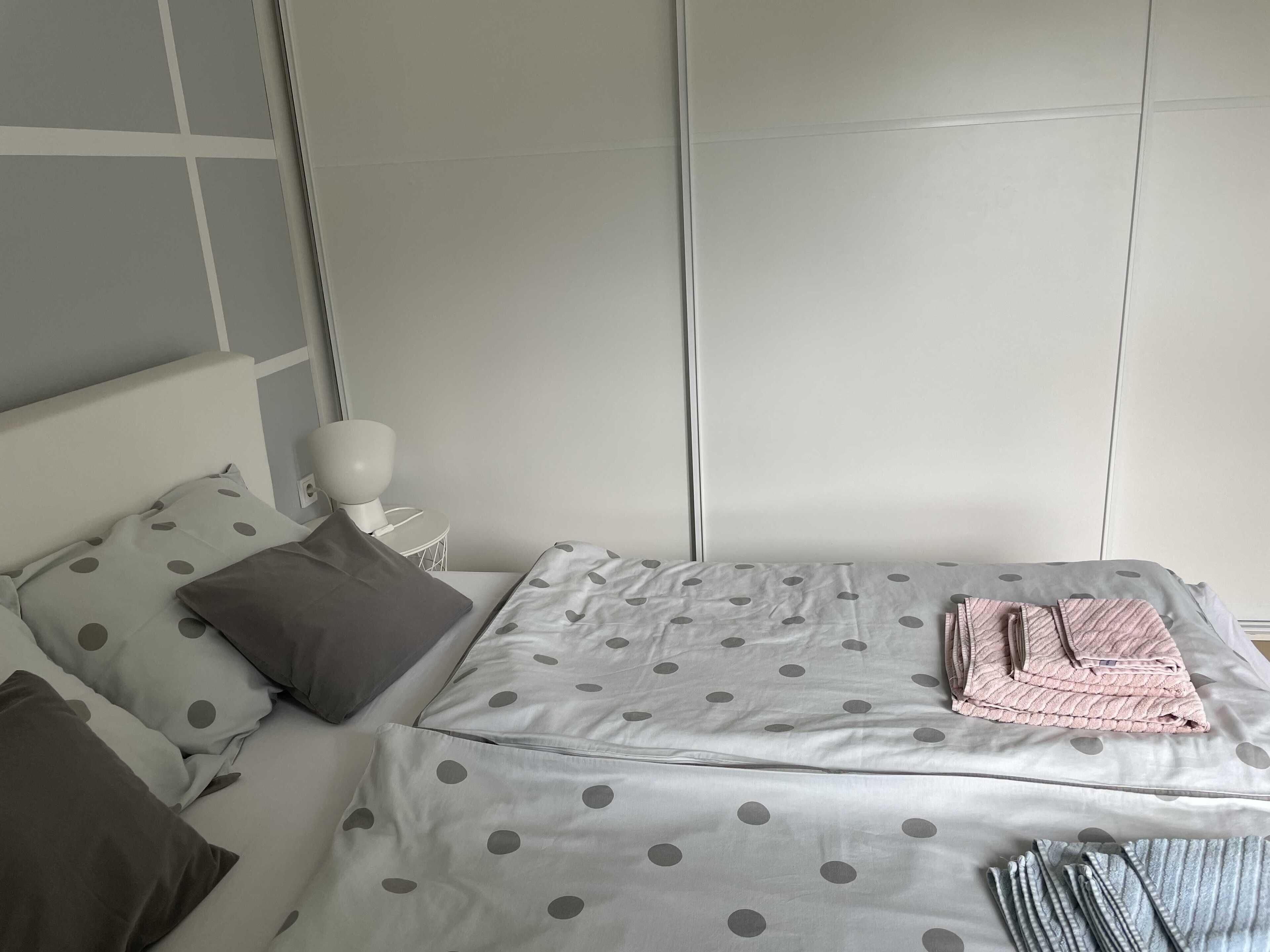 Un lit bien fait, avec une couvre-lit à pois et des oreillers, une table de chevet avec une lampe, et un mur sobre.