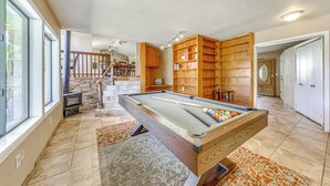 Una mesa de billar en una habitación con chimenea de piedra, estanterías de madera y una escalera.