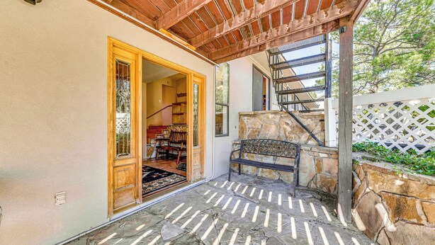 Un patio de piedra con una pérgola de madera, un banco de piedra y una escalera que conduce a un nivel superior.