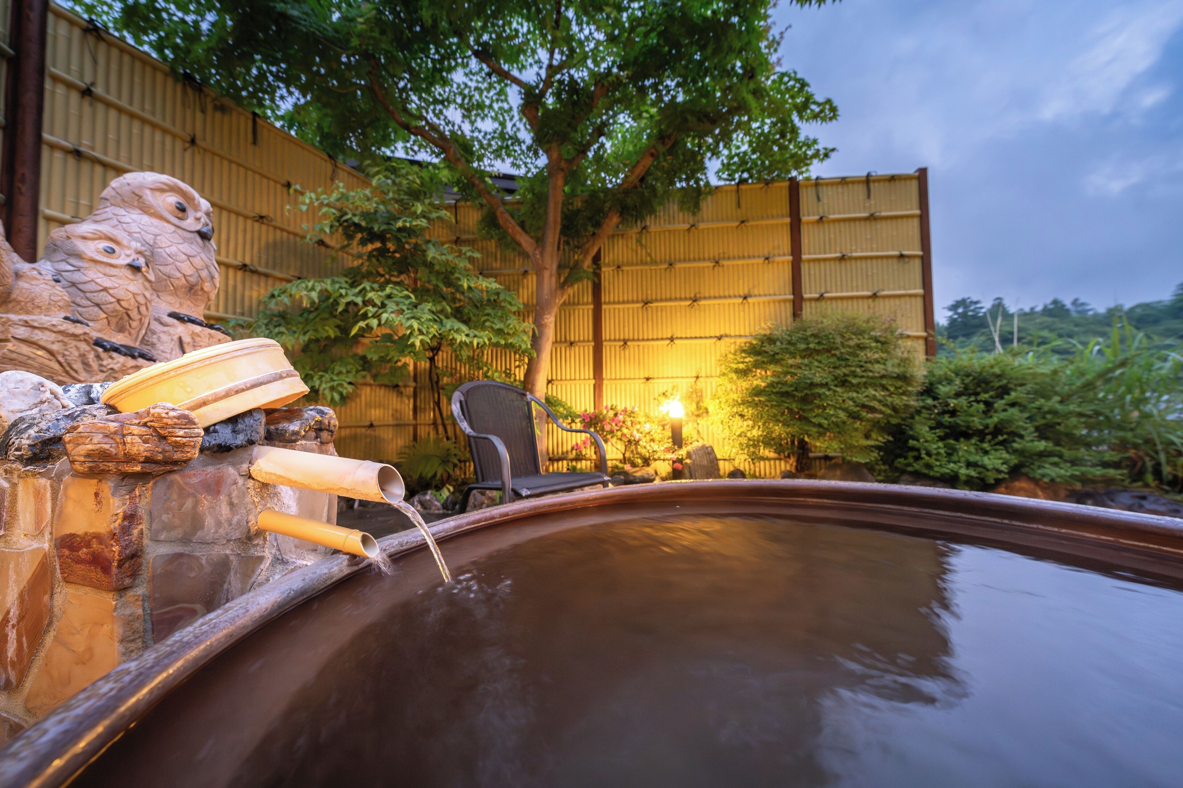 A hot spring area with stone sculptures, a wooden bench, and a bamboo fence in the background.