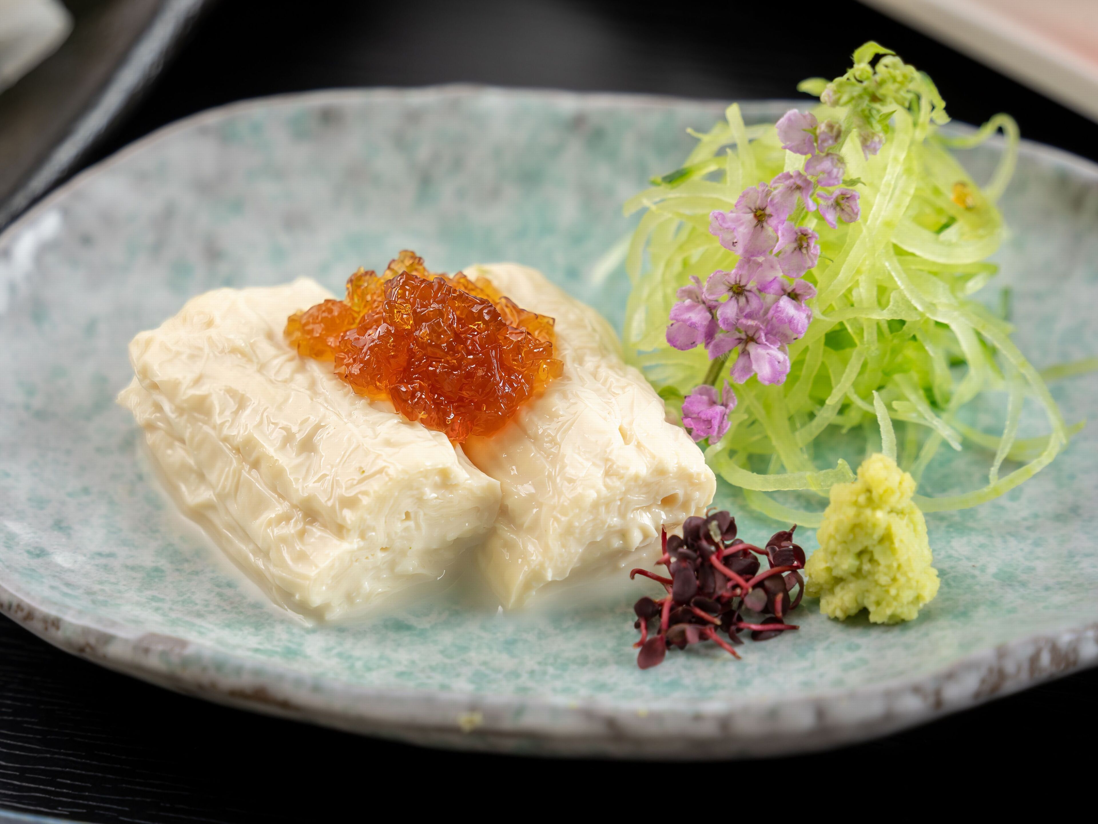 Um prato com um creme coberto por caviar laranja, acompanhado por uma salada de legumes verdes e flores roxas.