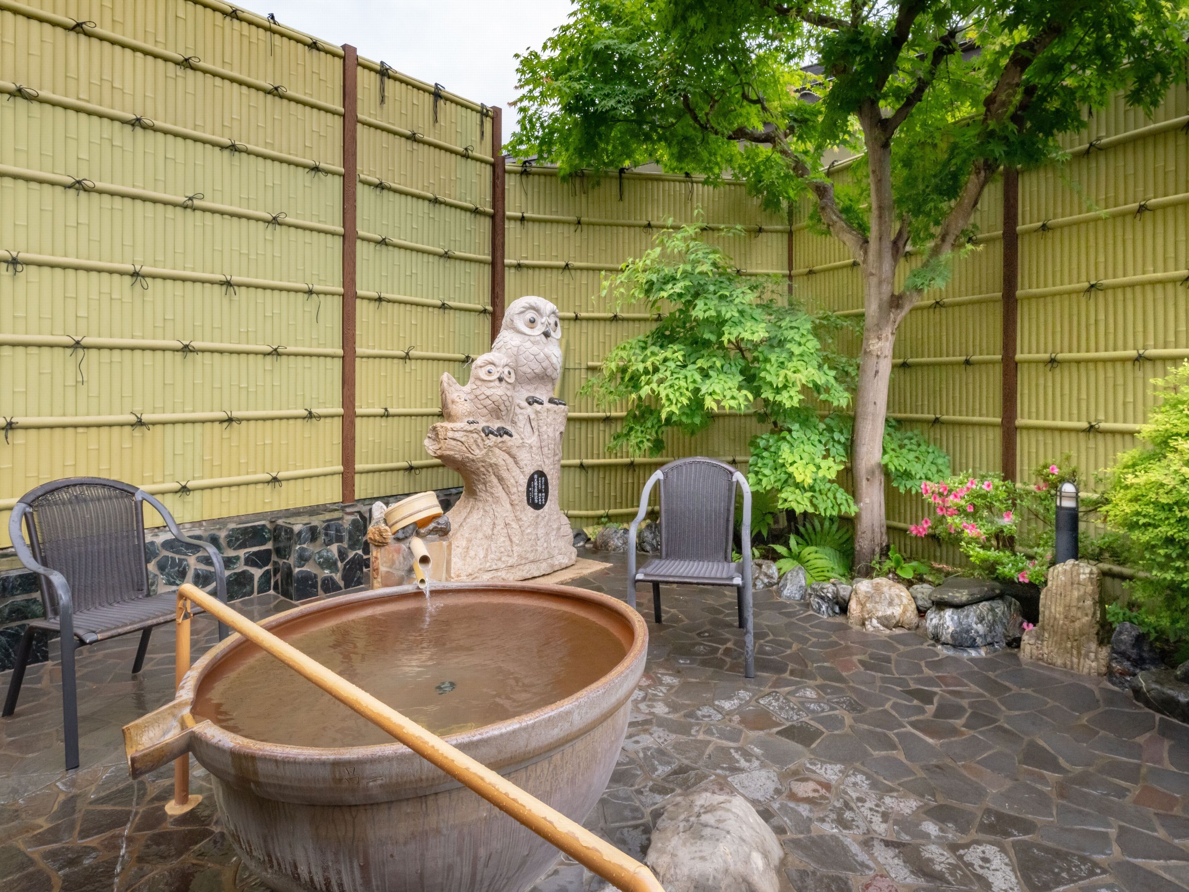 Un jardí japonès tradicional amb una font termal de pedra, dues cadires de vimetes, una taula de pedra i una tanca de bambú.
