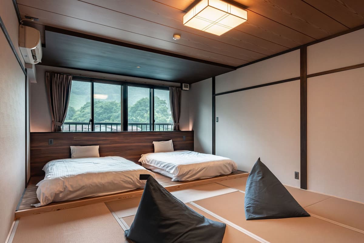 A traditional Japanese-style room with tatami flooring, two beds, a view of greenery, and a sliding door.