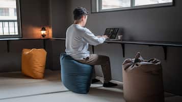 En person sidder på en blå pølle-stol ved et skrivebord med en bærbar computer, i et rum med et stort vindue, en gulvlampe og en brun pølle på gulvet.