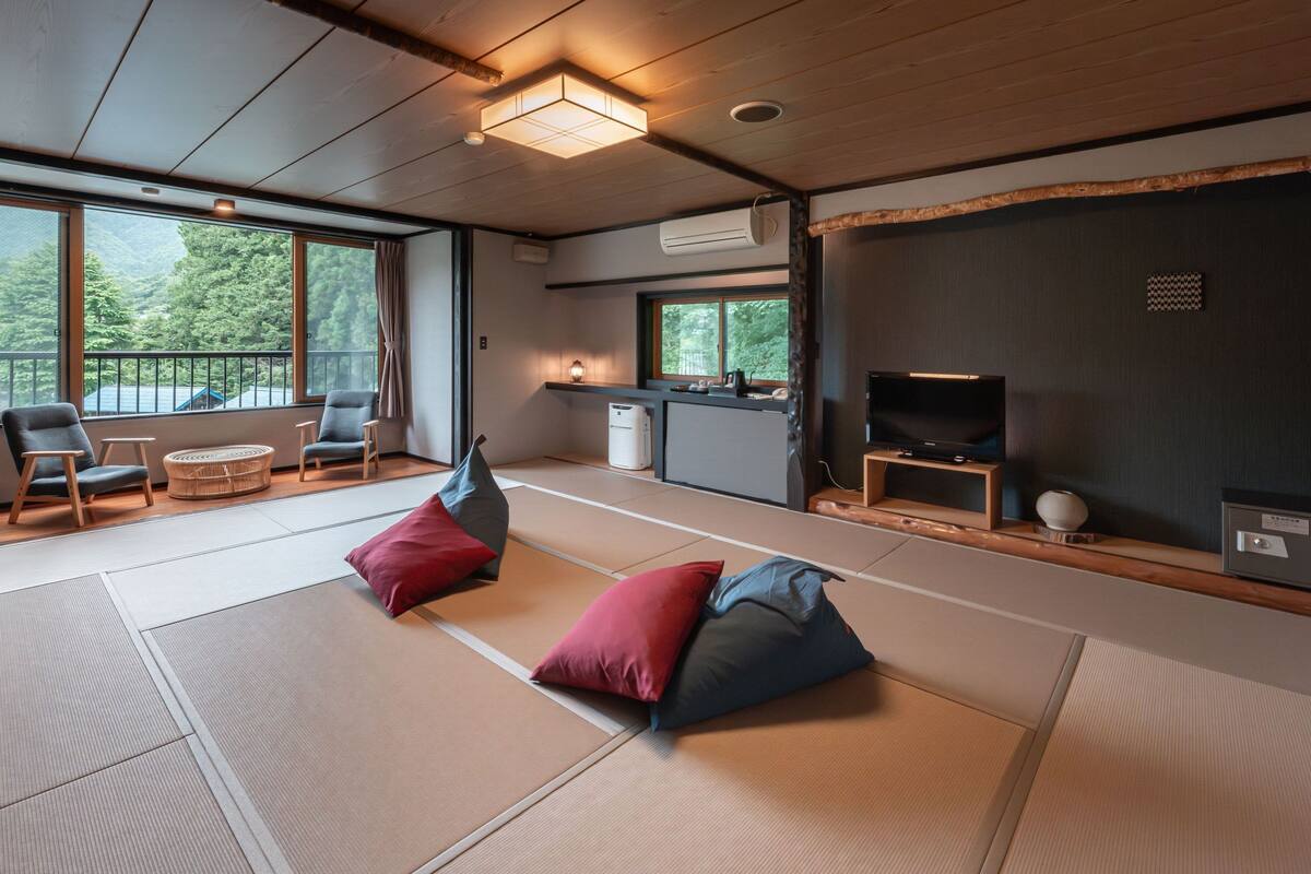 A modern room with tatami mats, a TV, a small kitchen, and large windows overlooking greenery.