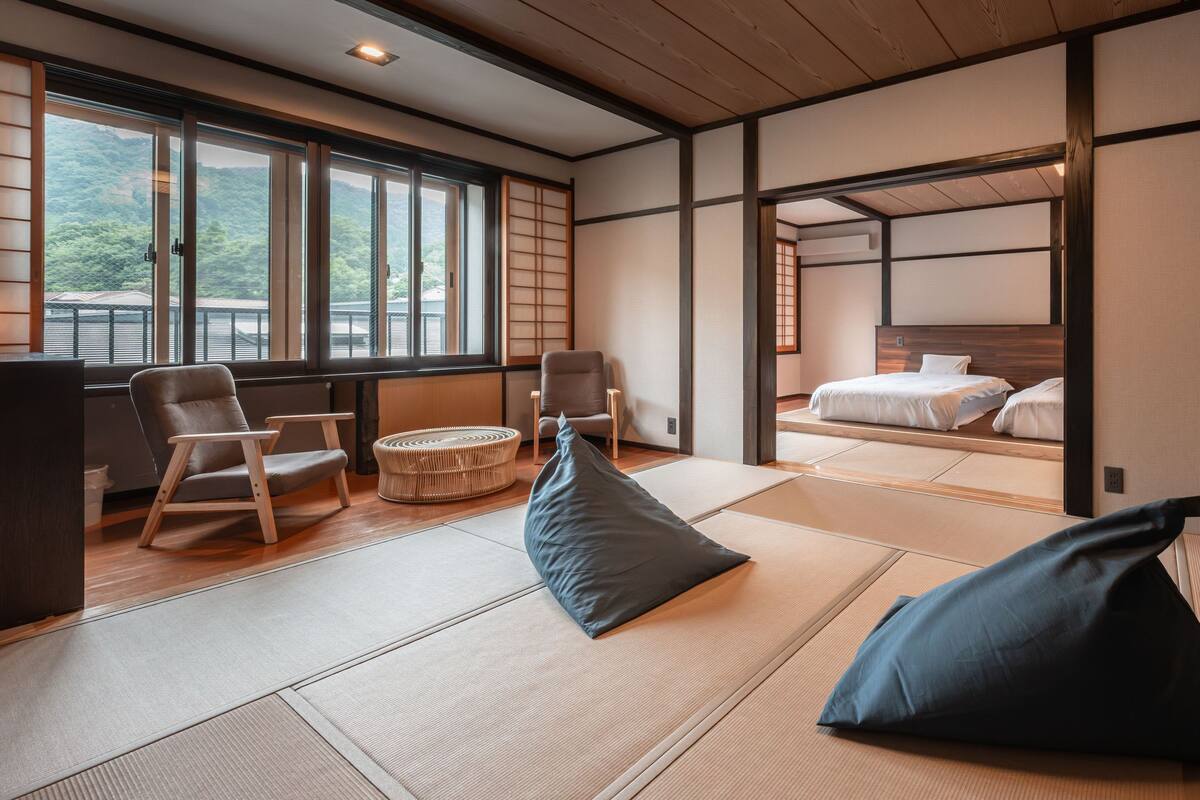 A traditional Japanese room with tatami flooring, a low wooden bed, and a view of a forested area through large windows.
