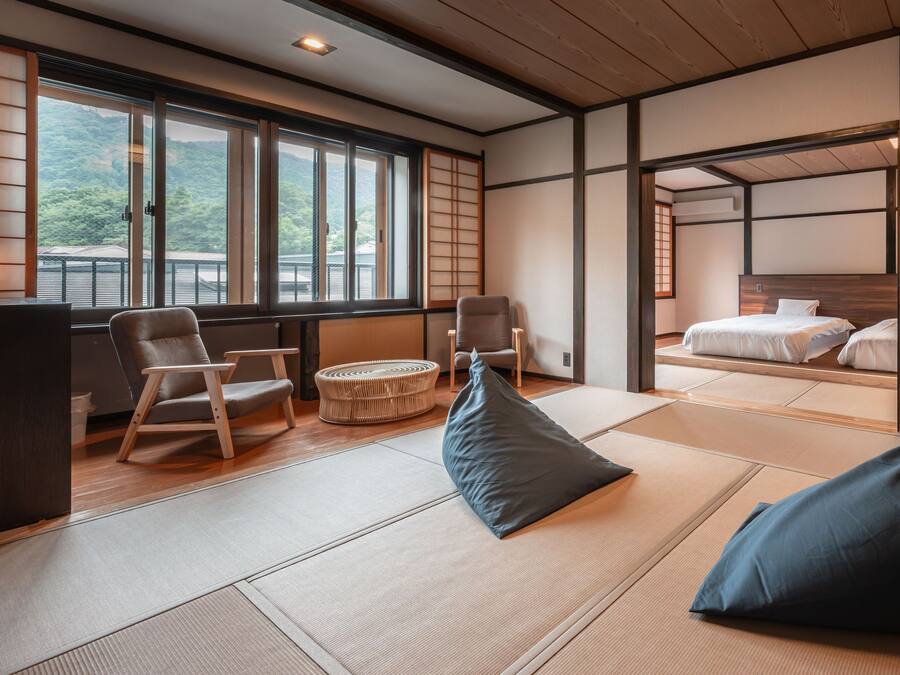 Una habitación japonesa tradicional con suelo de tatami, una cama de madera baja y vistas a una zona boscosa a través de amplios ventanales.