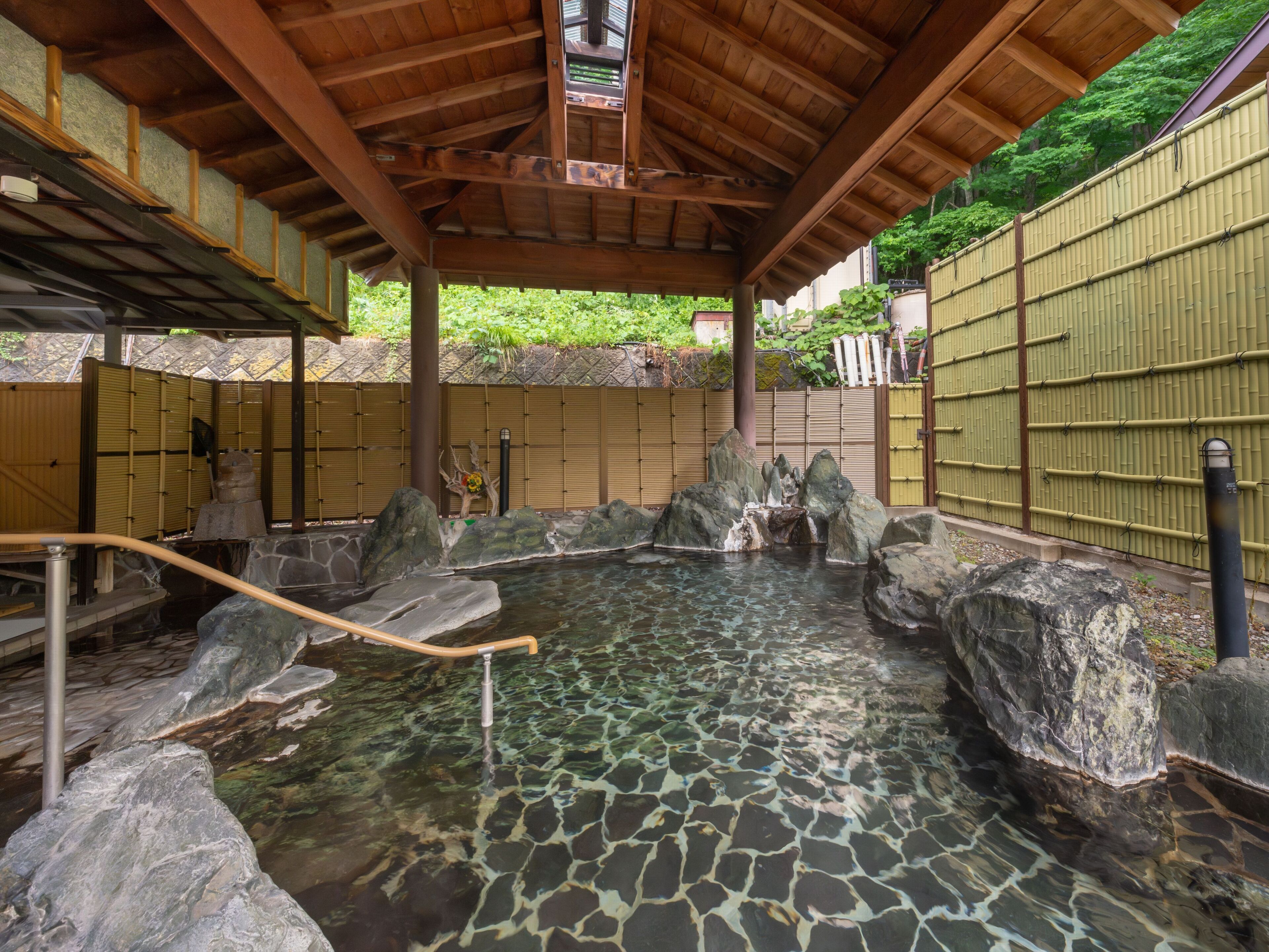 A traditional Japanese onsen with a wooden roof, stone basin, and bamboo walls.