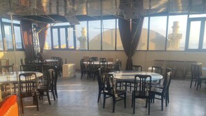 A spacious dining area with multiple round tables and chairs, large windows with curtains, and a view of distant structures. - Duc Chung Hotel Ha Long (Bai Chay)