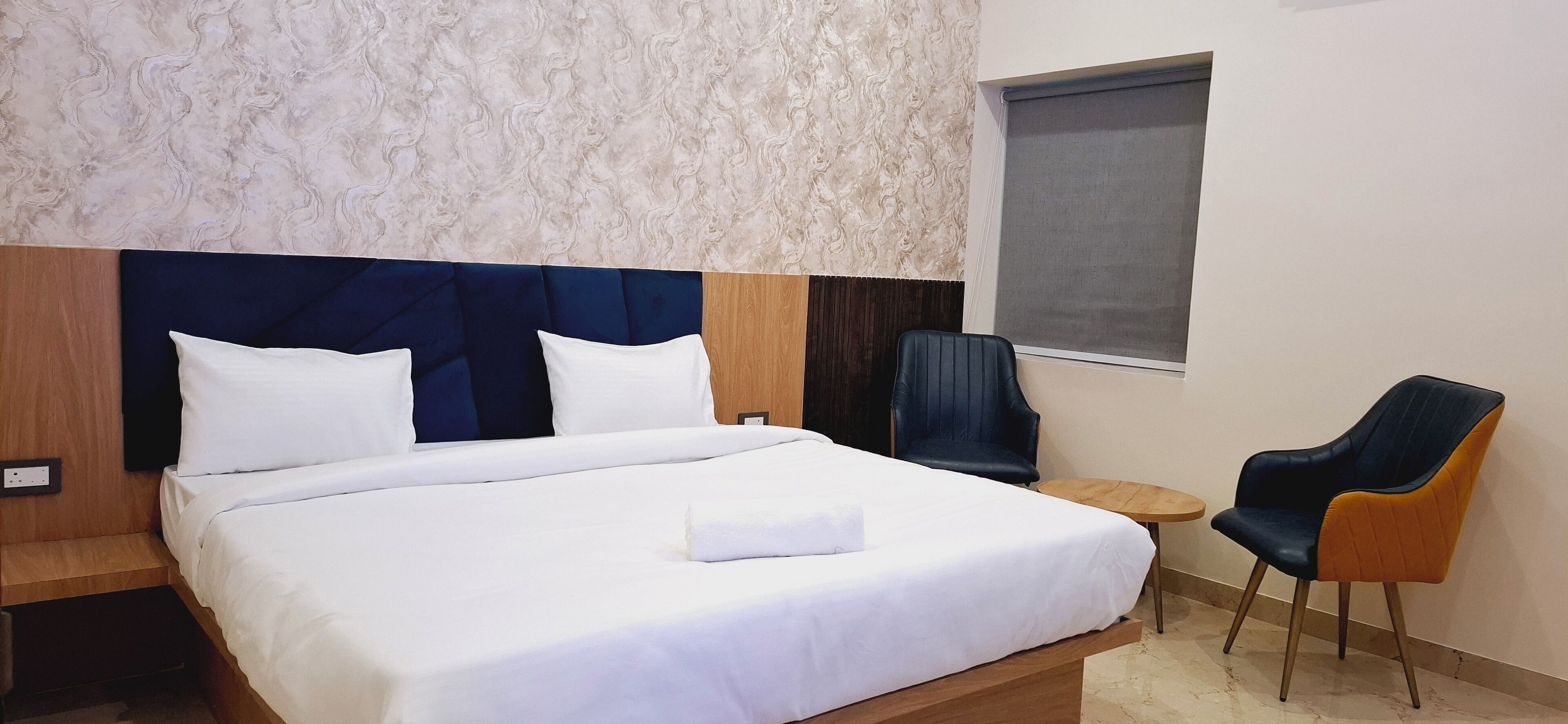 Une chambre d’hôtel avec un grand lit, deux oreillers, une tête de lit en bois, une petite table ronde, une chaise bleue et une fenêtre avec un store.