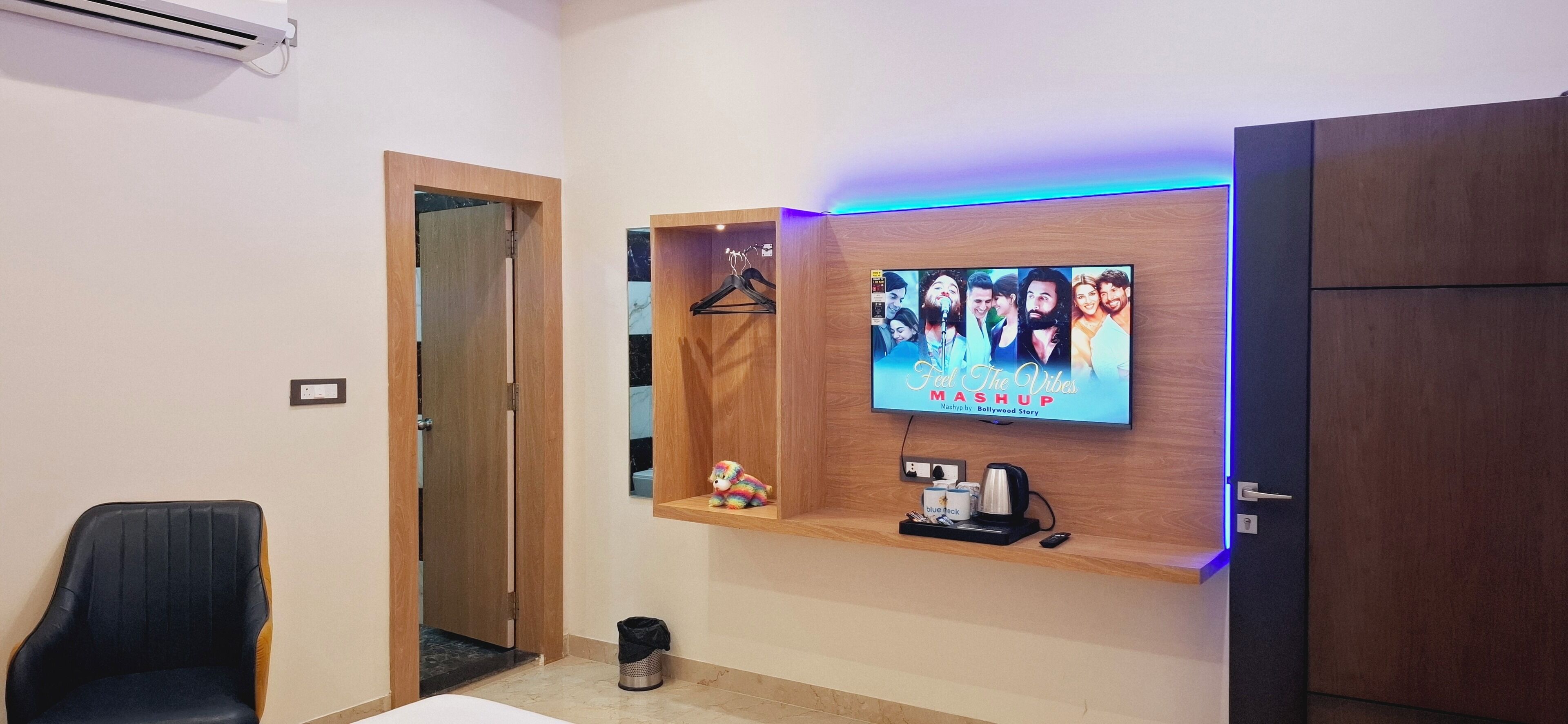 Hotellihuoneessa on seinälle asennettu televisio, jolla näkyy Bollywood Storyn musiikkikappale ”Feel The Vibes Mashup”. Television alla on puinen hylly, jonka päällä on kahvinkeitin, muki ja pieni koriste-esine. Musta tuoli on sijoitettu television vasemmalle puolelle.