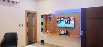 A hotel room with a TV mounted on the wall displaying 'Feel The Vibes Mashup' by Bollywood Story. There is a wooden shelf below the TV holding a coffee machine, a mug, and a small figurine. A black chair is positioned to the left of the TV.