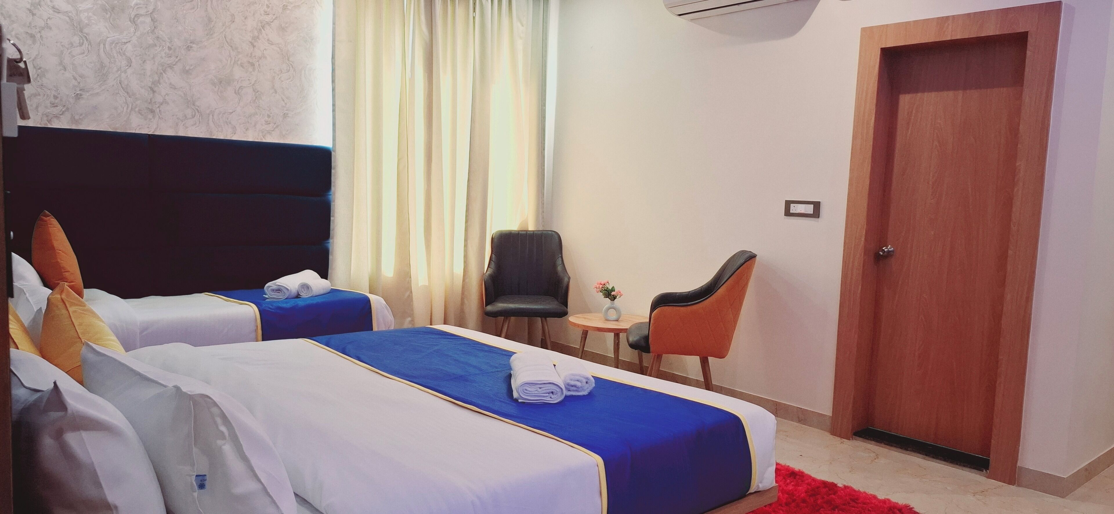 A hotel room with two beds, a wooden door, a small table with a vase, and two chairs.