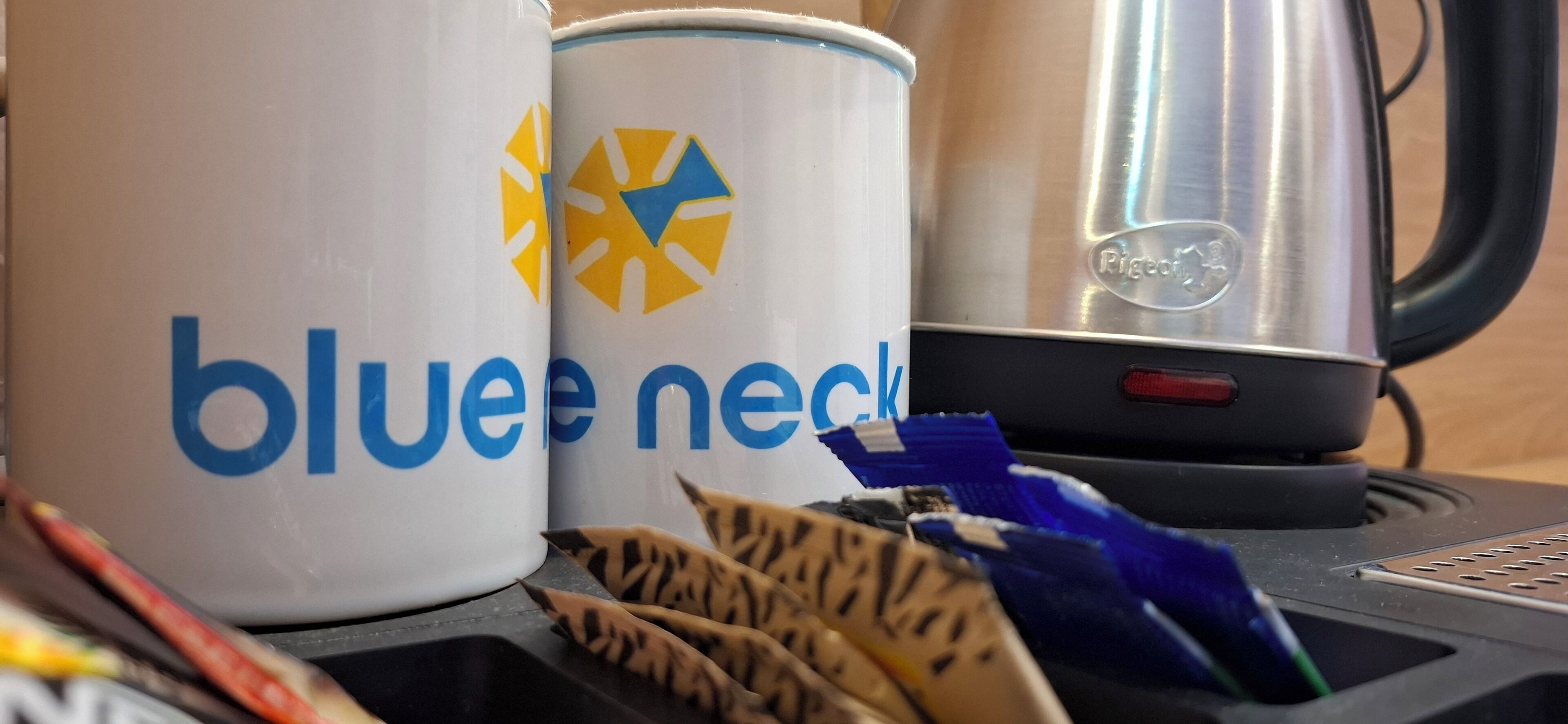 Two white mugs with blue text and a logo, a stainless steel kettle, and a black coffee machine on a countertop.