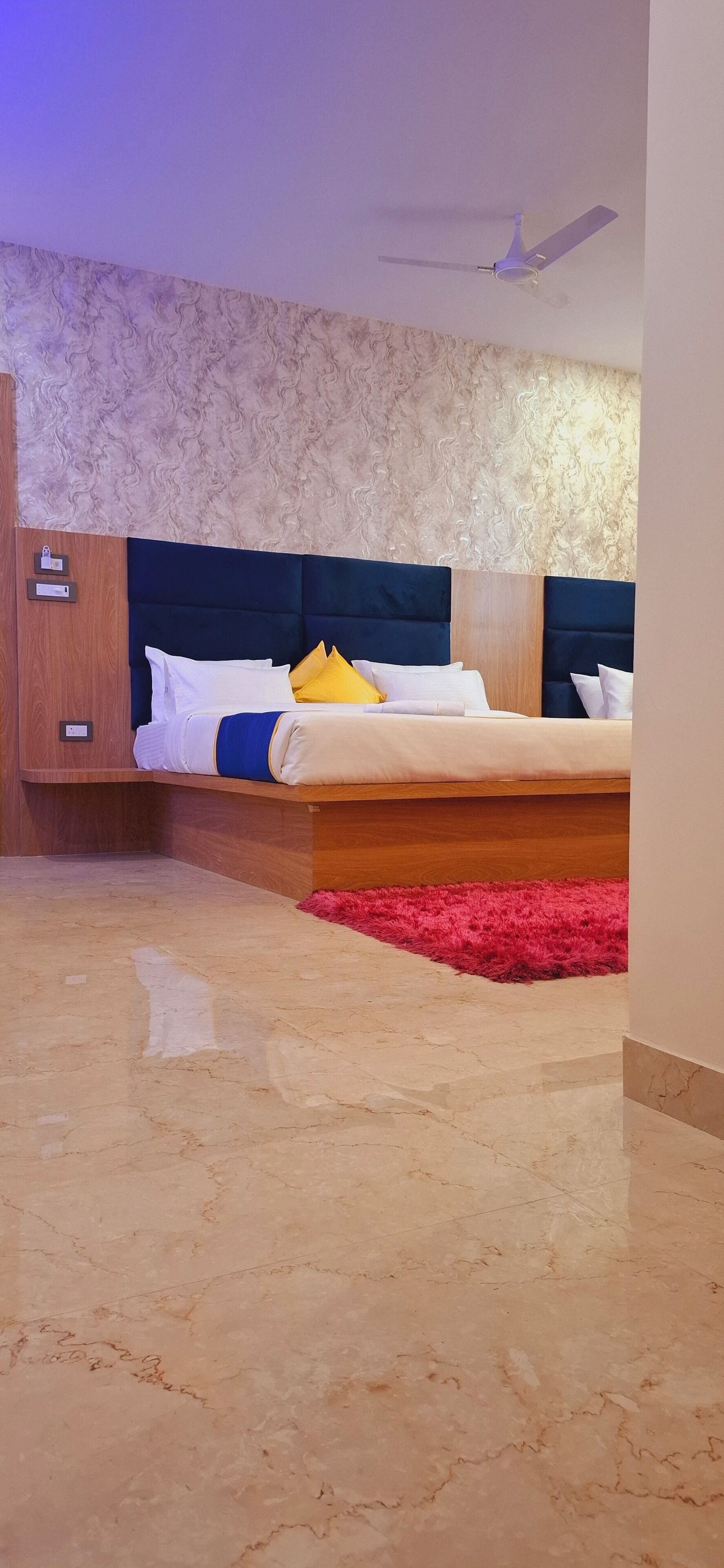 A modern hotel room with a marble floor, a bed with white and yellow bedding, a wooden wardrobe, and a ceiling fan.