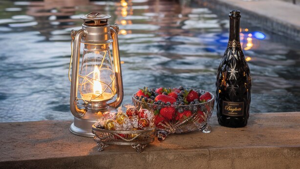 Escena junto a la piscina con un farol, una botella y un cuenco de fruta.