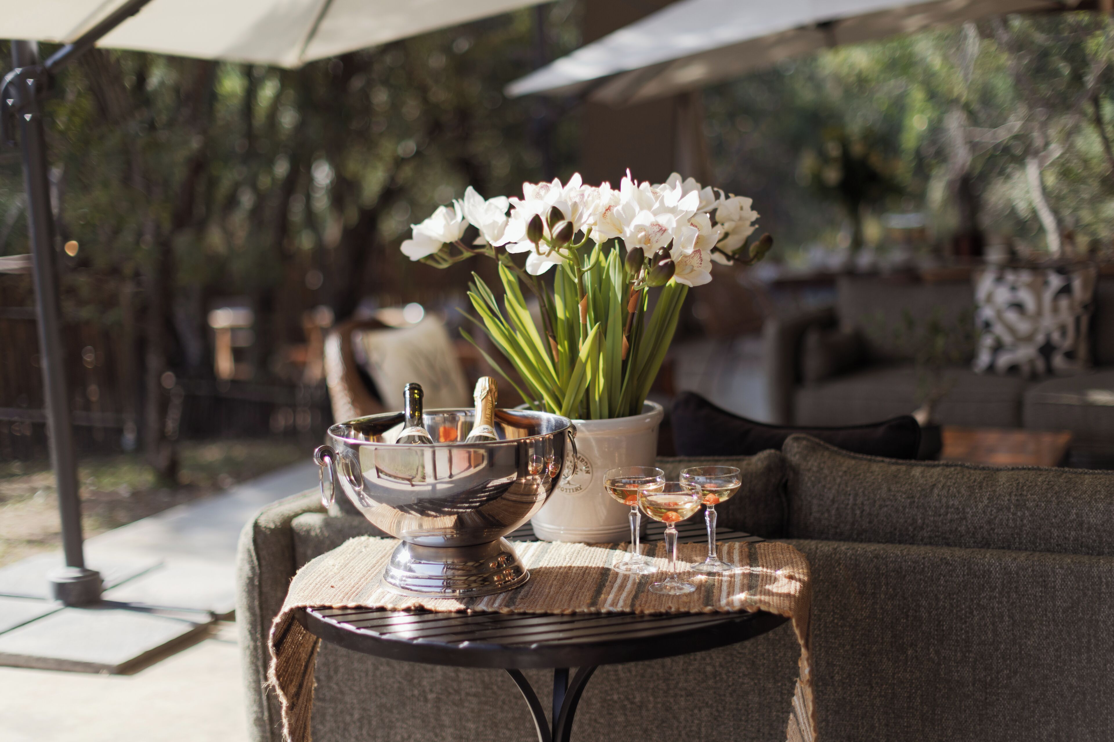 A table with a metal bowl, a vase with flowers, and two glasses of a clear liquid, possibly wine, on a textured tablecloth.