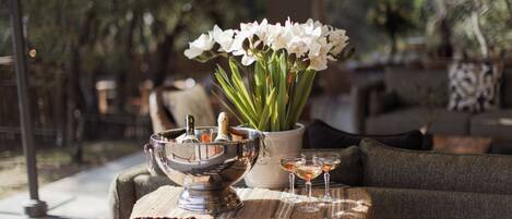 A table with a metal bowl, a vase with flowers, and two glasses of a clear liquid, possibly wine, on a textured tablecloth.