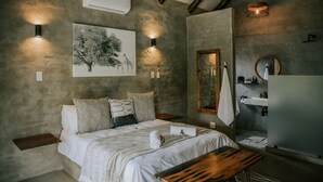 Un dormitorio con cama, mesitas de noche, aire acondicionado en la pared, lámpara colgante, espejo y baño con lavabo y toallero.