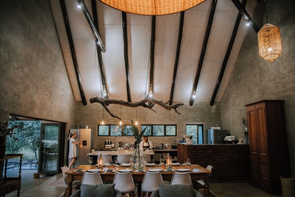 A dining area with a long table set for multiple guests, a chef preparing food, and a rustic ceiling with exposed beams and a hanging light fixture. - Rhino's Rest Private Luxury Villa (Hoedspruit)