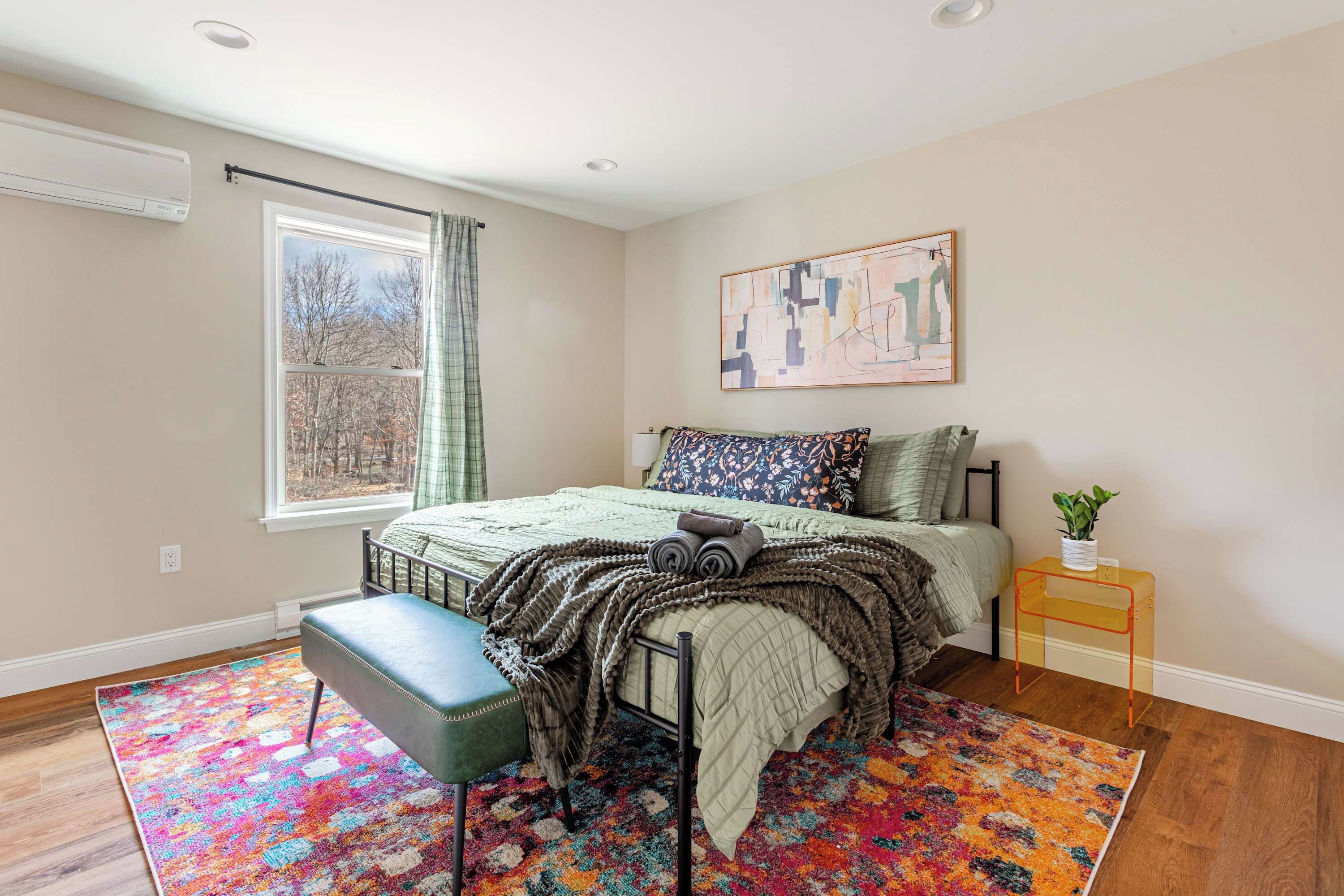A bedroom with a bed, a bench, a window with curtains, a painting on the wall, and a small table with a plant.