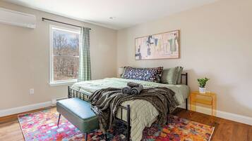 A bedroom with a bed, a bench, a window with curtains, a painting on the wall, and a small table with a plant.