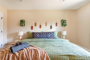 Cama bem arrumada com colcha verde, dois travesseiros e um cobertor dobrado. Há duas luminárias de mesa e uma planta na parede.