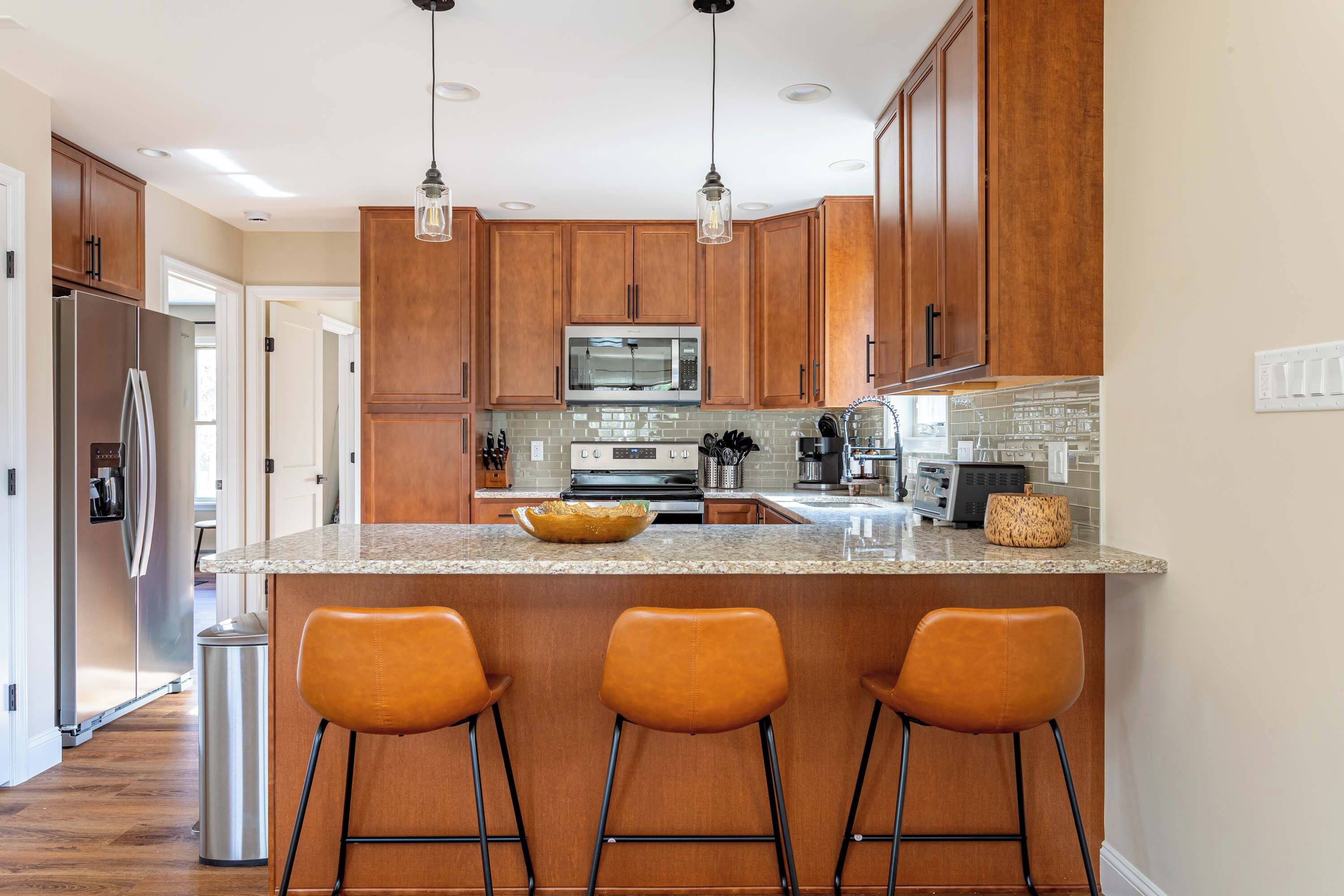 A kitchen with wooden cabinets, a stainless steel refrigerator, a microwave, and a granite countertop. There are three orange bar stools and a trash can.