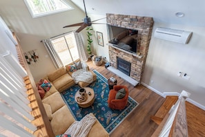 A spacious living room with a fireplace, a ceiling fan, and a large window.