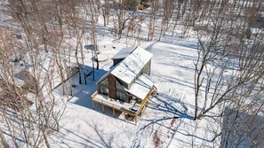 A snow-covered cabin with a deck and a large window, surrounded by bare trees.
