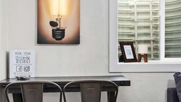 Een eetgedeelte met een tafel en stoelen, een wandlamp, een lijst met een afbeelding en een raam met jaloezieën.