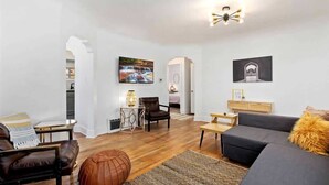 Ein modernes Wohnzimmer mit Sofa, Sessel und Hocker, einem Couchtisch und einem Fernseher. Ein Kamin, eine Lampe und Kunstwerke an den Wänden sind ebenfalls vorhanden.