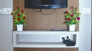 A flat-screen TV mounted on a wooden panel wall, flanked by two white shelves with red and pink flowers. - Hotel Delhi Darshan -By RSL Hospitality (New Delhi)