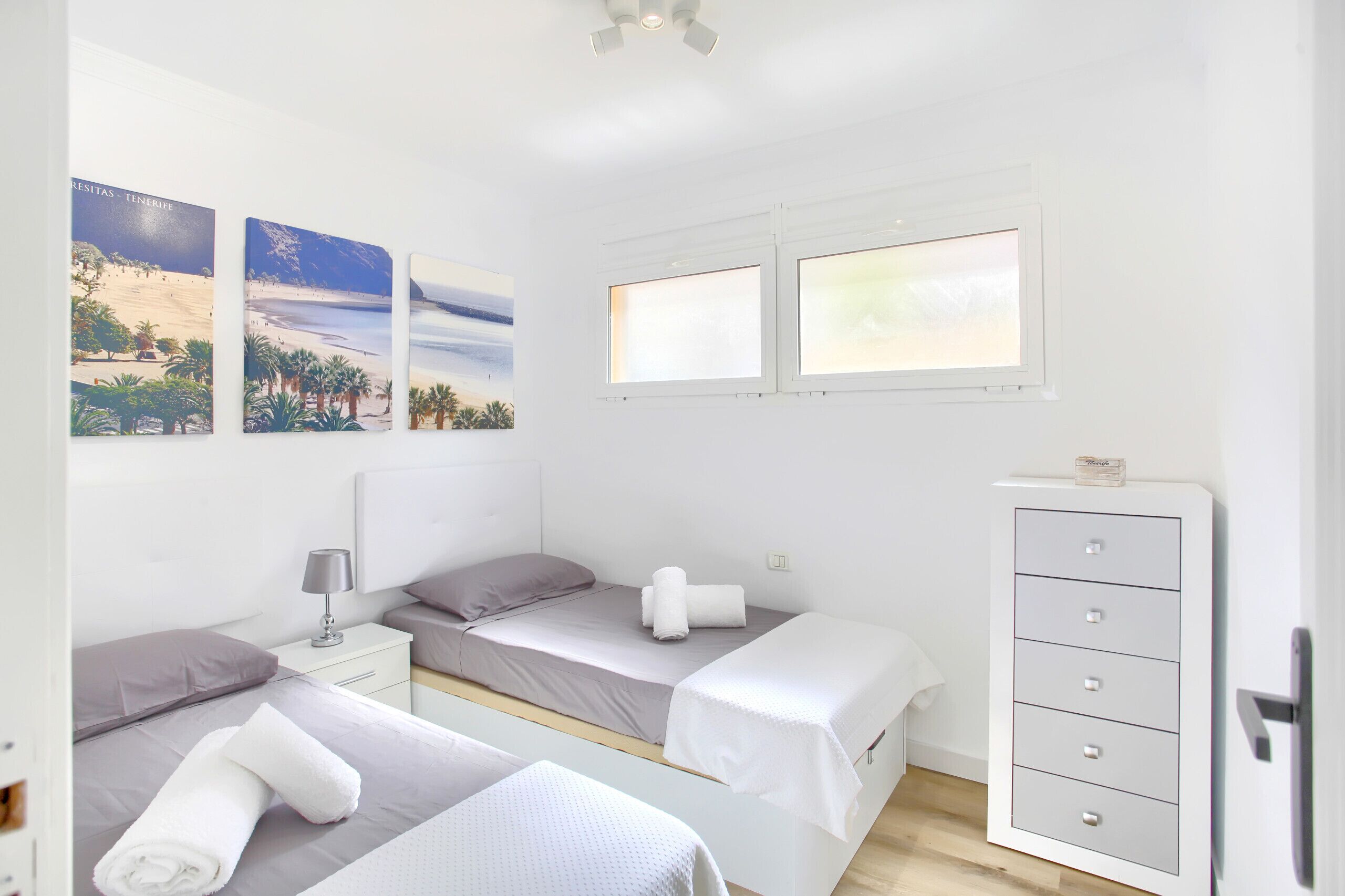 Une chambre avec deux lits simples, une table de chevet avec une lampe, une commode et trois tableaux encadrés accrochés au mur.