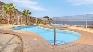A swimming pool with a curved design, surrounded by a fence, palm trees, and a mountainous backdrop. - The Penthouse, Fabulous Sunset, Heated Pool, Fiber (Costa Adeje)