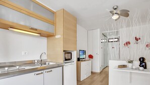 A modern kitchen with built-in appliances, a microwave, and a sink. There is a white cabinet with drawers and a wooden cabinet above the microwave. A ceiling fan is visible, and a decorative wall with white branches is present. - Ocean Touch Studio, Pool, Wi-Fi, Near the Beach (Playa de Las Americas)