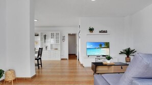 A modern living room with a grey sofa, a flat-screen TV, and a wooden coffee table. There is a dining area with a black chair and a white cabinet.