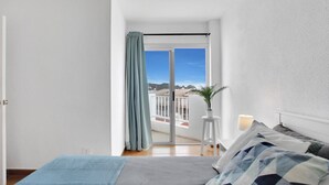 A bedroom with a bed, a nightstand, a potted plant, and a view of a balcony with a railing.